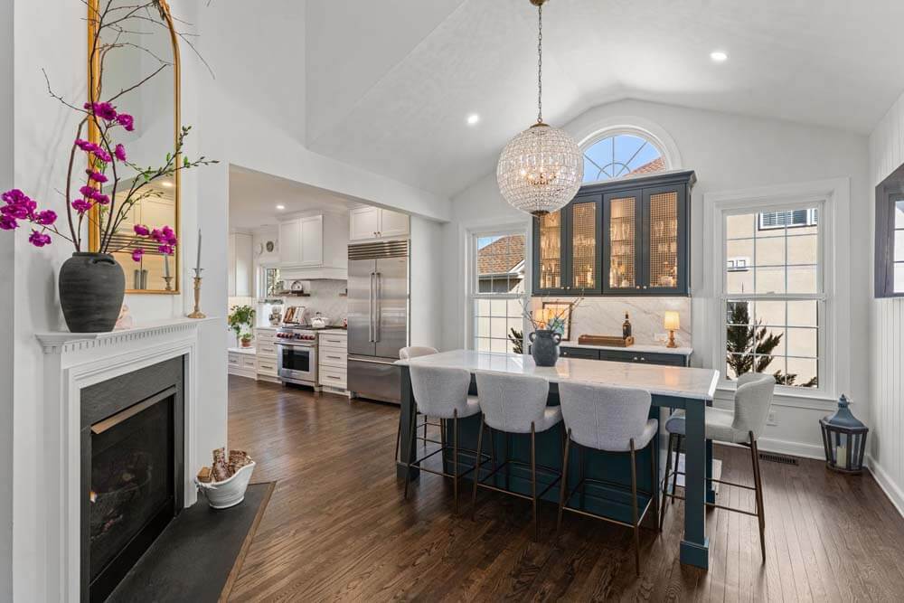 Bright open-concept kitchen and dining area with a marble island, six gray stools, and a crystal chandelier overhead.