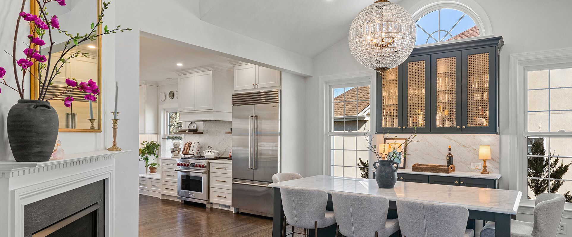 Bright open kitchen with marble island, white cabinetry, stainless refrigerator, and a large crystal chandelier over a dark-blue display cabinet.